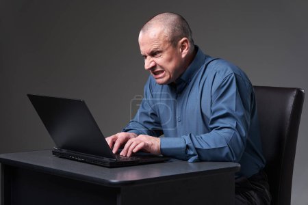 Téléchargez les photos : Frustré homme d'affaires mature tapant furieusement à son ordinateur portable sur son bureau - en image libre de droit