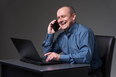 Photo for Mature businessman working at his desk on a laptop and talking at the cellphone in the same time - Royalty Free Image