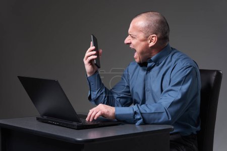 Photo for Very angry and demanding mature businessman yelling in a phone conference, while being at his laptop - Royalty Free Image