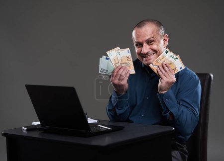 Téléchargez les photos : Heureux homme d'affaires mature tenant de l'argent, assis à son bureau, image conceptuelle de la richesse, bon revenu, transactions - en image libre de droit