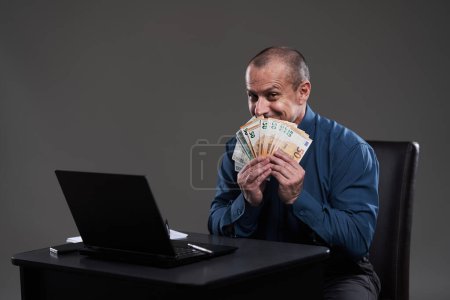 Téléchargez les photos : Heureux homme d'affaires mature tenant de l'argent, assis à son bureau, image conceptuelle de la richesse, bon revenu, transactions - en image libre de droit