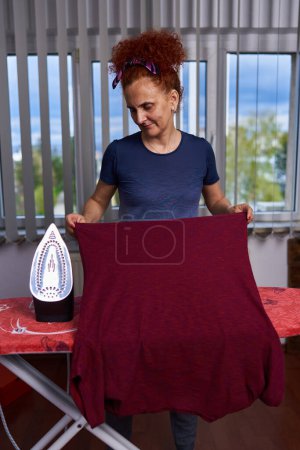 Téléchargez les photos : Cheveux bouclés rousse femme au foyer repassage vêtements par la fenêtre - en image libre de droit