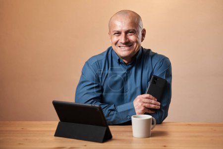 Foto de Feliz hombre de negocios maduro sentado en su escritorio, con un teléfono y una tableta y una taza de café, sobre fondo cervatillo - Imagen libre de derechos
