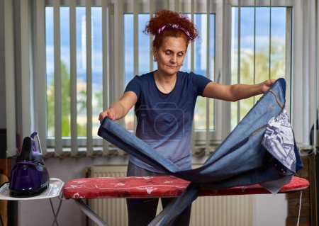 Téléchargez les photos : Cheveux bouclés rousse femme au foyer repassage vêtements par la fenêtre - en image libre de droit
