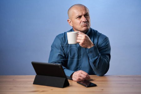 Photo for Pensive mature business man having coffee at his desk, in front of his tablet - Royalty Free Image