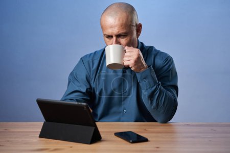 Photo for Pensive mature business man having coffee at his desk, in front of his tablet - Royalty Free Image