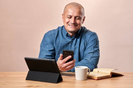 Photo for Happy mature business man sitting at his desk, having a phone and a tablet and a cup of coffee, over fawn background - Royalty Free Image