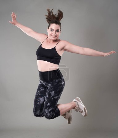Photo for Young confident plus size woman in gym clothing jumping high, on gray background - Royalty Free Image