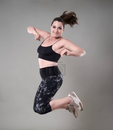 Téléchargez les photos : Jeune femme confiante plus la taille dans les vêtements de gymnastique sautant haut, sur fond gris - en image libre de droit