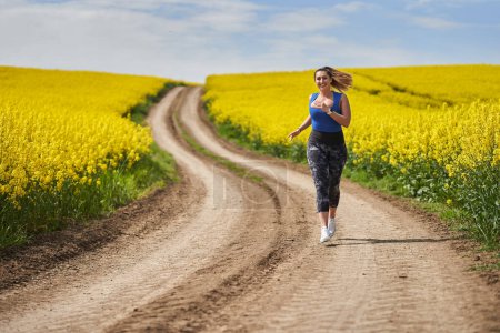 Photo for Plus size beautiful latin woman jogging on a dirt road by a canola field - Royalty Free Image