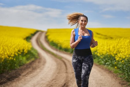 Téléchargez les photos : Plus la taille belle femme latine jogging sur un chemin de terre par un champ de canola - en image libre de droit