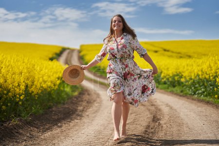 Téléchargez les photos : Plus la taille magnifique femme dans une robe florale, courir et danser pieds nus sur un chemin de terre par un champ de canola florissant dans la campagne - en image libre de droit