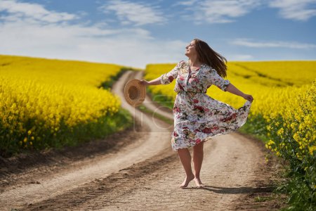 Photo for Plus size gorgeous woman in a floral dress, running and dancing barefoot on a dirt road by a blossoming canola field in the countryside - Royalty Free Image