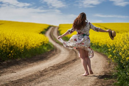 Photo for Plus size gorgeous woman in a floral dress, running and dancing barefoot on a dirt road by a blossoming canola field in the countryside - Royalty Free Image