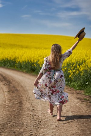Photo for Romantic beautiful blonde hispanic young woman in a floral dress, walking barefoot on a dirt road going through a canola field - Royalty Free Image
