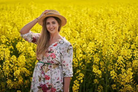 Photo for Portrait of a beautiful woman farmer in straw hat and floral dress by a flowering canola field - Royalty Free Image