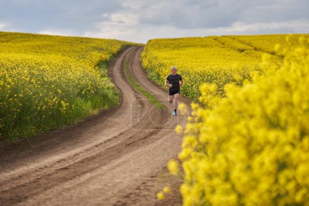 Téléchargez les photos : Coureur de distance d'âge moyen courant sur un chemin de terre dans un champ de canola en fleurs - en image libre de droit