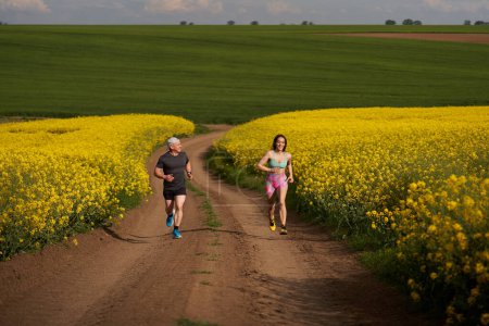 Foto de Un par de corredores corriendo en un curso montañoso en una pista de tierra en un campo de canola - Imagen libre de derechos