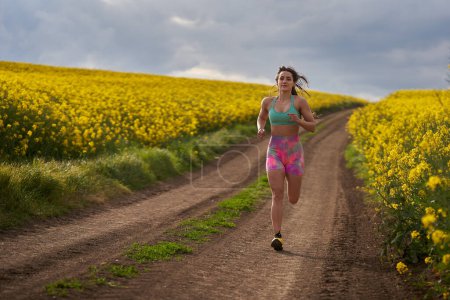 Photo for Young woman runner running on a dirt track through a blooming canola field - Royalty Free Image