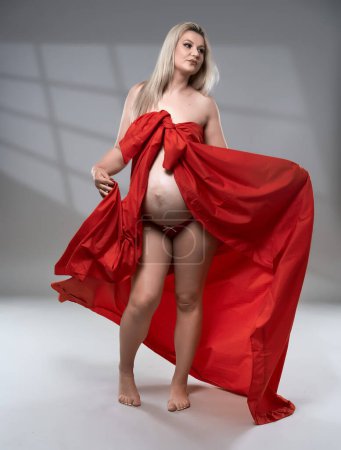 Photo for Beautiful pregnant young blonde woman in red wavy sheet on gray background - Royalty Free Image