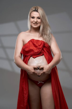 Téléchargez les photos : Belle jeune femme blonde enceinte en feuille ondulée rouge sur fond gris - en image libre de droit