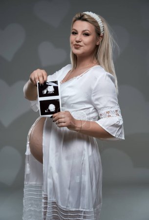 Téléchargez les photos : Belle jeune femme blonde enceinte en robe blanche sur fond gris - en image libre de droit