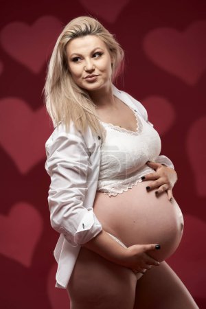 Foto de Hermosa embarazada joven rubia en lencería blanca sobre fondo rojo - Imagen libre de derechos