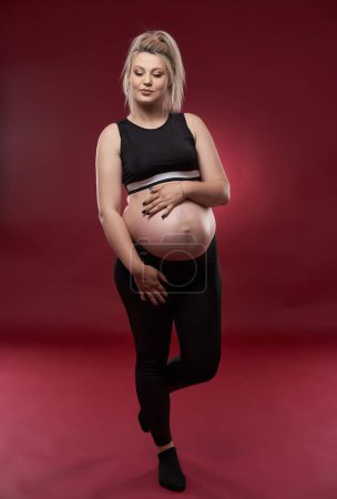 Photo for Beautiful pregnant young blonde woman in black clothes, studio shot on red background - Royalty Free Image