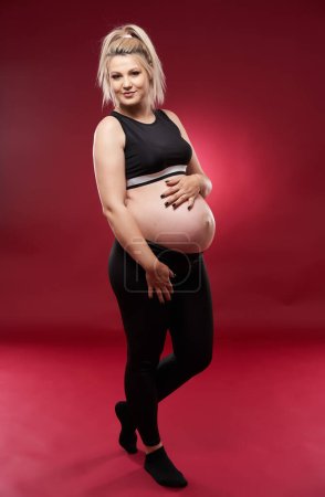 Téléchargez les photos : Belle jeune femme blonde enceinte en vêtements noirs, studio tourné sur fond rouge - en image libre de droit