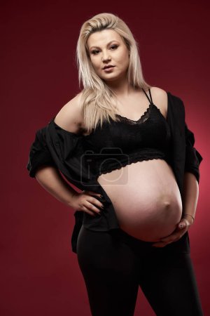 Photo for Beautiful pregnant young blonde woman in black clothes, studio shot on red background - Royalty Free Image