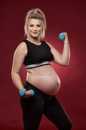 Young pregnant woman doing fitness exercises on red background, studio shot 
