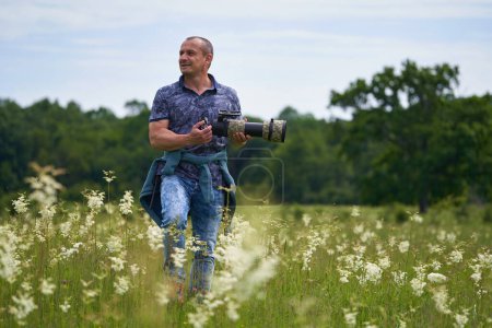 Téléchargez les photos : Photographe de nature professionnel sur le terrain près de la forêt, tenant un appareil photo avec un téléobjectif long - en image libre de droit