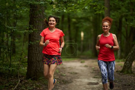 Foto de Dos mujeres felices y sanas corriendo por un sendero para correr en el bosque - Imagen libre de derechos