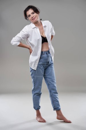 Foto de Atractiva mujer morena descalza en camisa blanca y jeans sobre fondo gris - Imagen libre de derechos