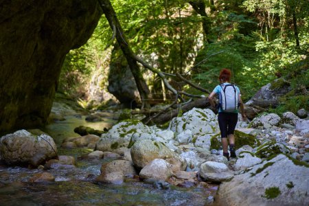 Foto de Mujer excursionista con mochila explorando gargantas salvajes junto a un río con rápidos y enormes rocas cubiertas de musgo - Imagen libre de derechos