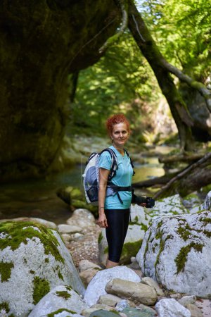 Foto de Mujer excursionista con mochila explorando gargantas salvajes junto a un río con rápidos y enormes rocas cubiertas de musgo - Imagen libre de derechos