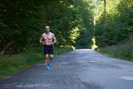Téléchargez les photos : Musclé athlétique homme torse nu courant sur une route asphaltée à travers la forêt - en image libre de droit