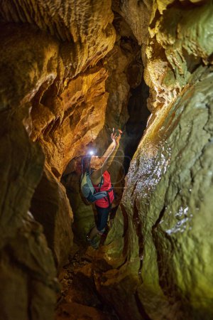 Foto de Mujer con mochila y linterna de cabeza explorando una cueva y tomando fotos - Imagen libre de derechos
