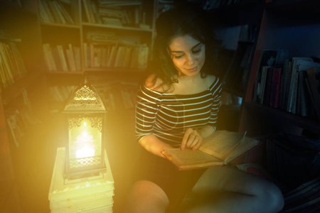 Photo for Beautiful brunette woman reading a book in a library at night, at the light of a lantern - Royalty Free Image