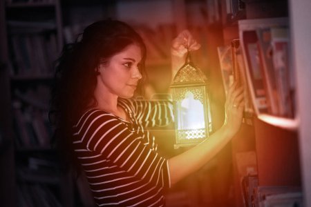 Téléchargez les photos : Belle femme brune lisant un livre dans une bibliothèque la nuit, à la lumière d'une lanterne - en image libre de droit