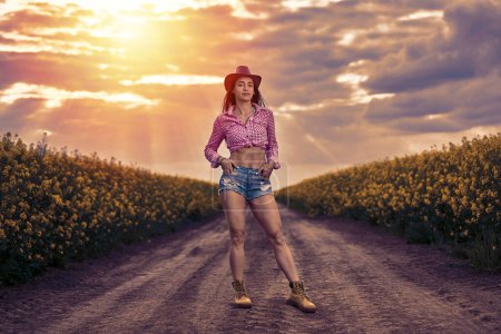 Foto de Atractiva campesina en pantalones cortos, camisa a cuadros y sombrero de pie en una carretera entre campos de canola al atardecer - Imagen libre de derechos