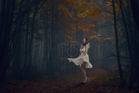 Téléchargez les photos : Belle jeune femme en robe blanche dansant dans une forêt mystérieuse aux couleurs automnales - en image libre de droit