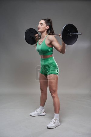 Photo for Young attractive woman fitness model working out with a heavy barbell, gray background - Royalty Free Image