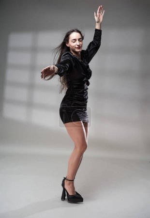 Foto de Mujer joven glamurosa en mini vestido negro con su pelo volando en el viento, estudio de tiro sobre fondo gris con la proyección de la ventana detrás - Imagen libre de derechos