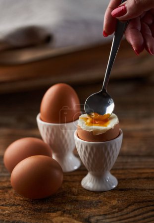Photo for Woman's hand with a spoon digging in a soft boiled egg in cup on a wooden board, runny yolk visible - Royalty Free Image