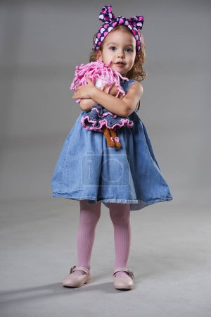Téléchargez les photos : Portrait d'une adorable petite fille blonde aux cheveux bouclés en robe bleue, tenant une poupée, sur fond gris, prise de vue studio - en image libre de droit