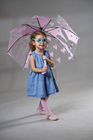 Foto de Retrato de una adorable niña rubia de pelo rizado con gafas enormes divertidas, sosteniendo un paraguas, sobre fondo gris, plano de estudio - Imagen libre de derechos