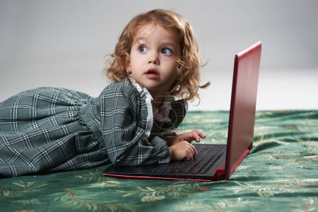 Téléchargez les photos : Portrait d'une adorable petite fille blonde aux cheveux bouclés jouant à des jeux sur un ordinateur portable, sur fond gris, plan studio - en image libre de droit