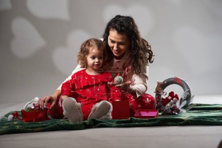 Téléchargez les photos : Petite fille tout-petit en pyjama de Noël jouant heureux avec sa mère, studio tourné sur fond gris - en image libre de droit