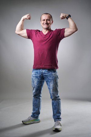 Téléchargez les photos : Homme d'âge moyen en tee et jeans fléchissant ses biceps sur fond gris - en image libre de droit
